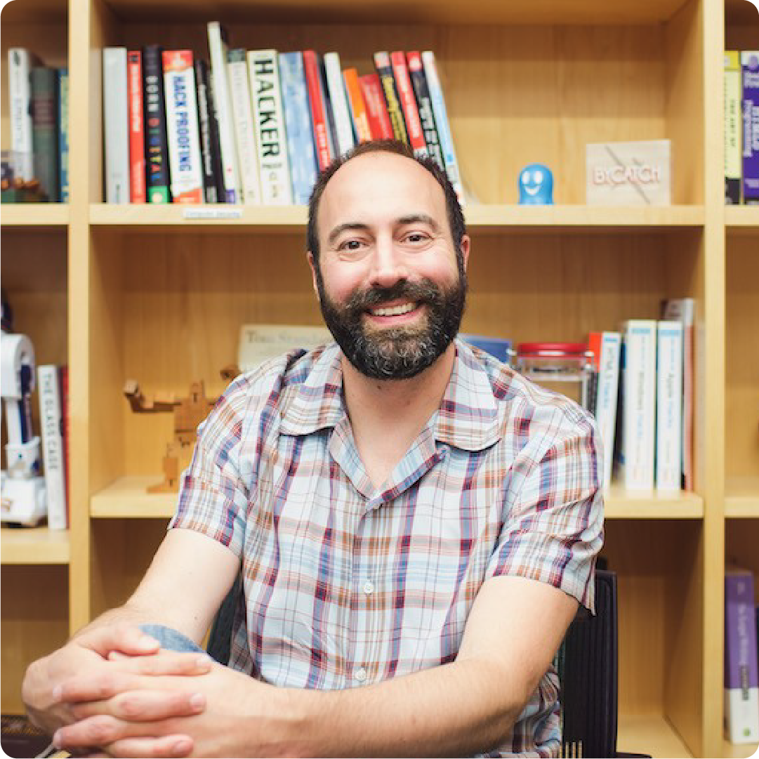A color photo of a man with a beard, sitting in front of a book shelf.