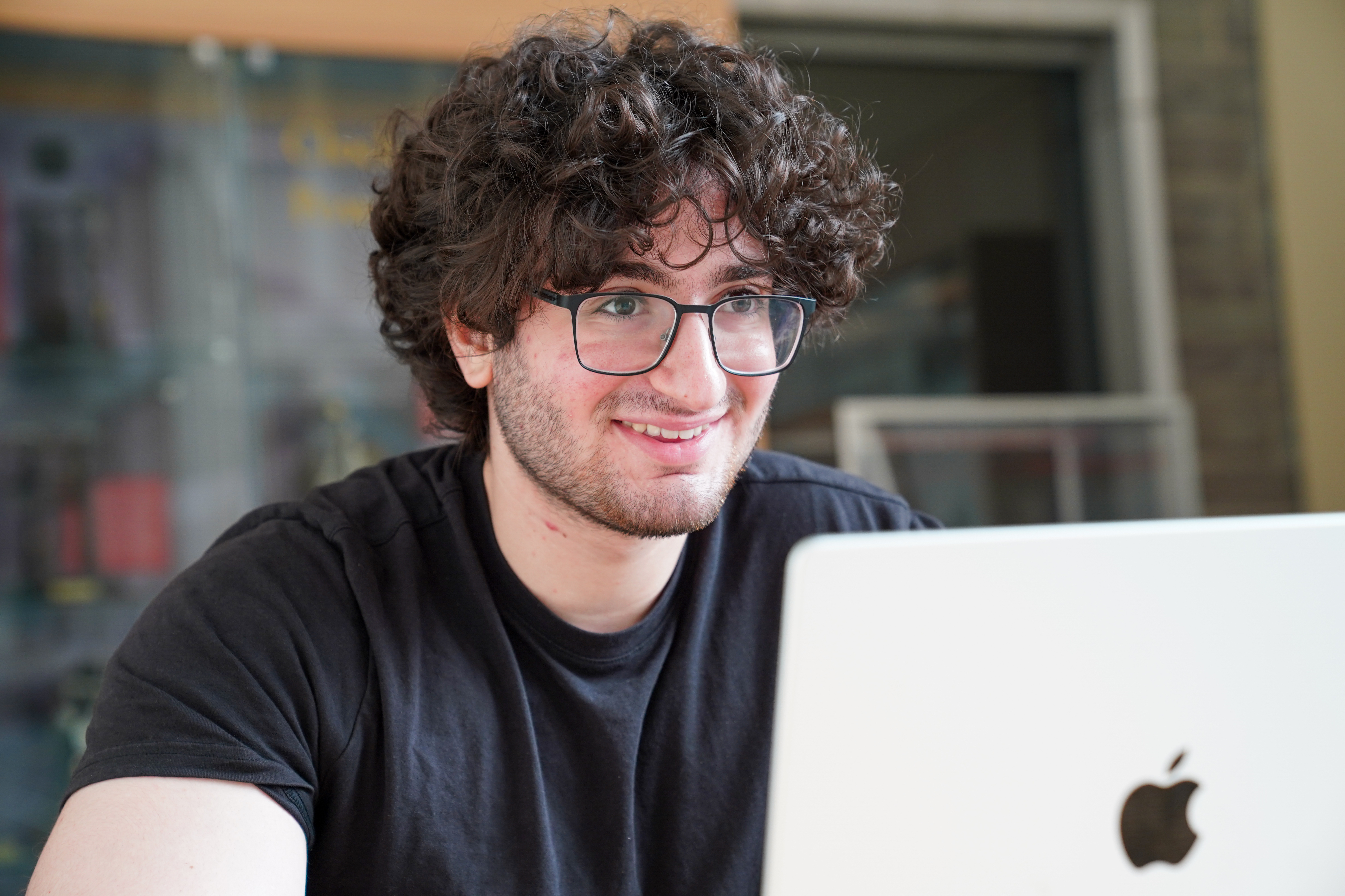 A color photo of a man with glasses, smiling while sitting beside his laptop.