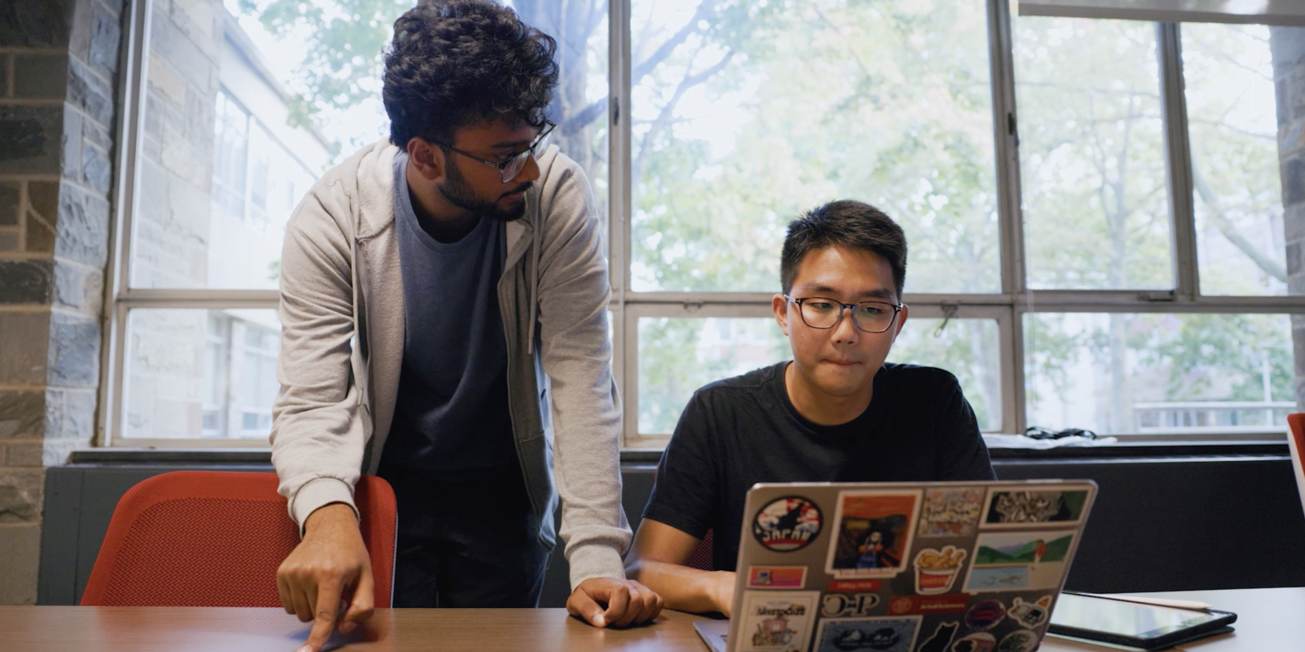two students (one standing, one sitting) look at computer laptop
