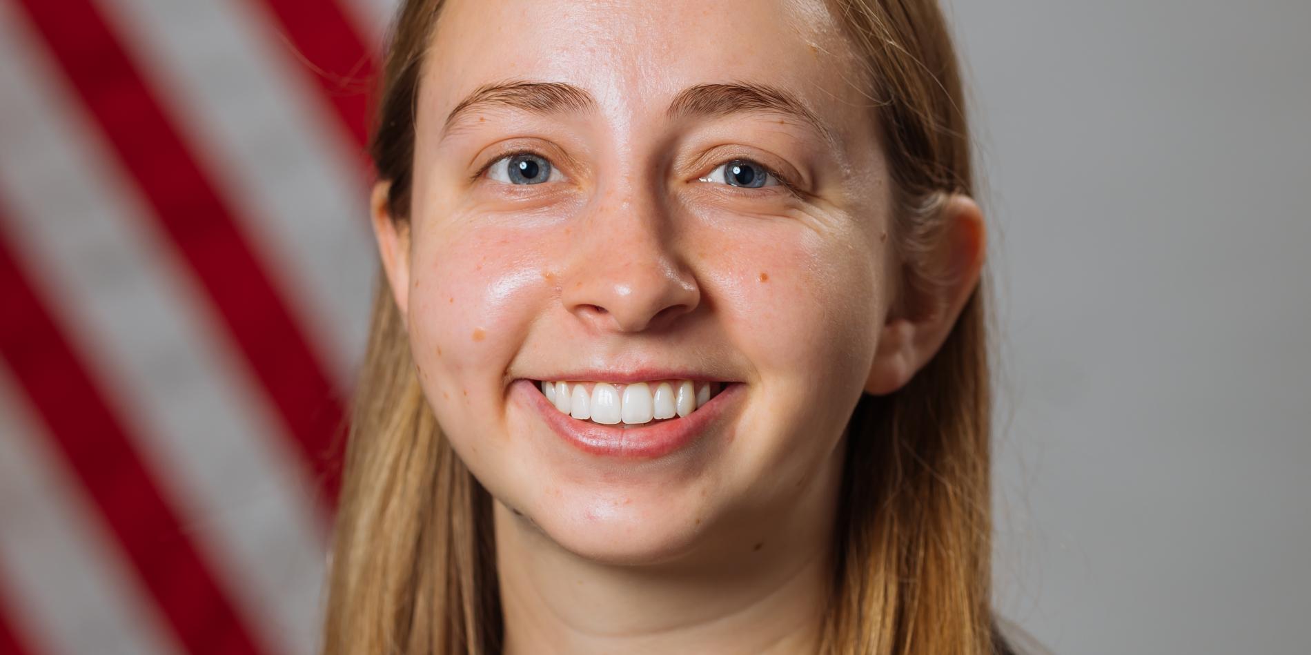 a female student with long blonde hair and a long sleeve black shirt sits in front of an American flag