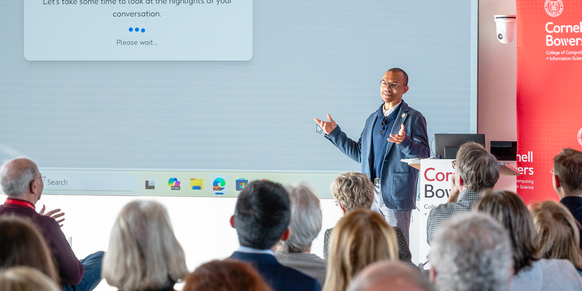 A color photo of a man giving a presentation in front of a large screen.