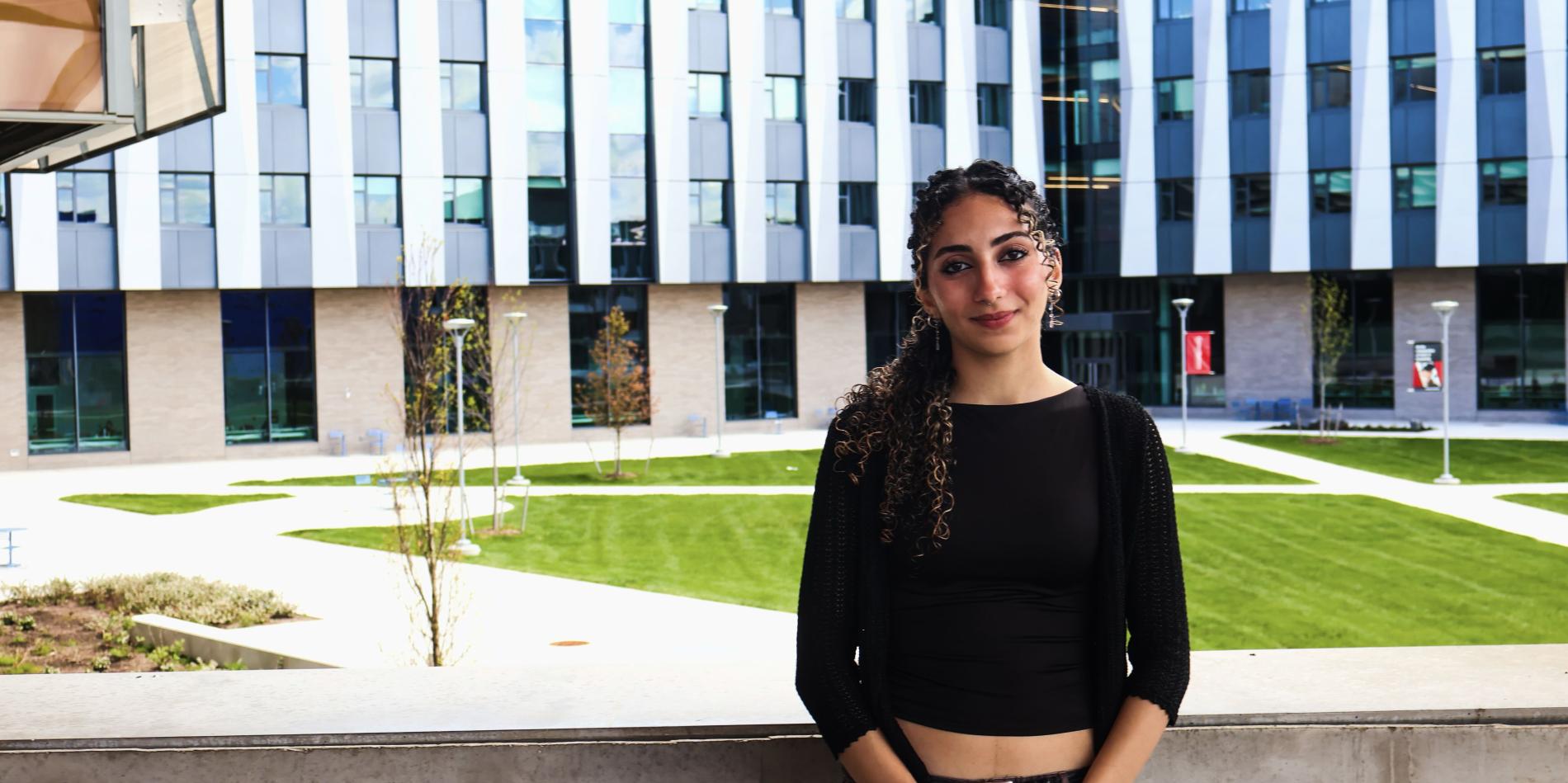 a young femal student with long brown hair wearing a black shirt and pants stands in front of a ledge with a large gray building in the background 