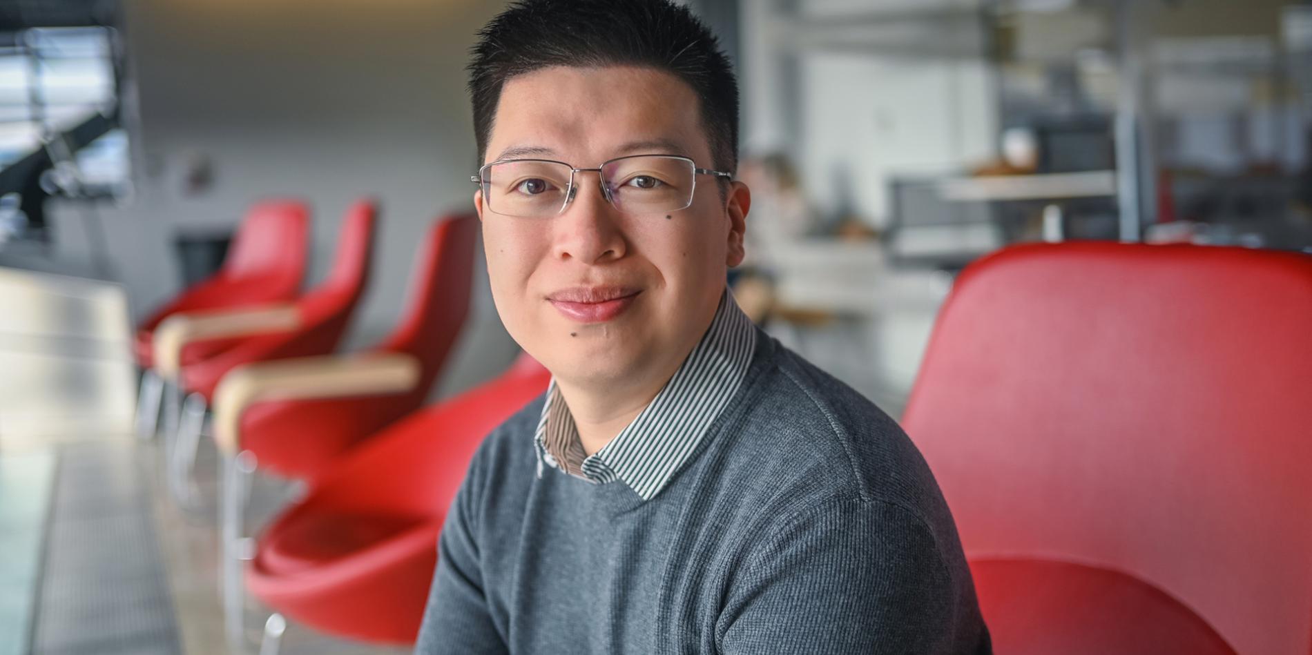 a male with dark brown hair and glasses waering a dark gray sweater with collar smiles as he sits on a red chair