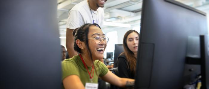 a woman sits at the computer smiling with two students in the background.