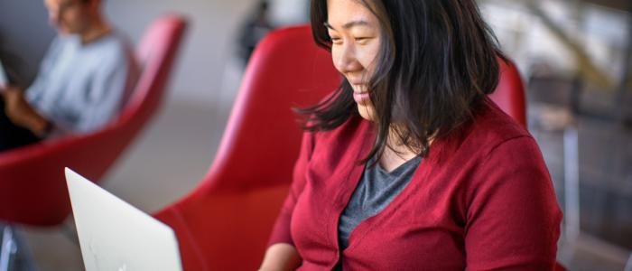 A woman sits in a red chair. She is smiling while working on a laptop and wearing a red cardigan.