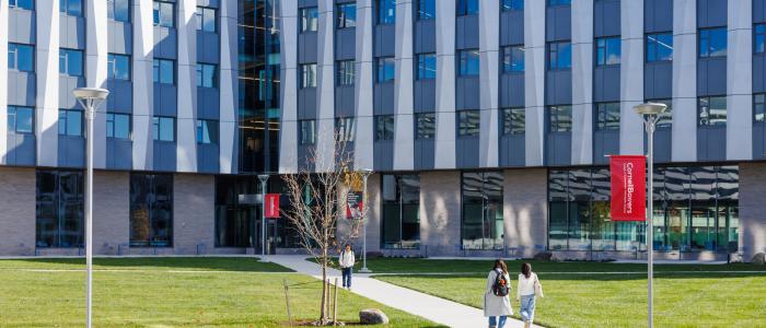 students walk along a paved path surrounded by green grass in front of a gray building