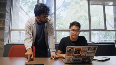 two students (one standing, one sitting) look at computer laptop
