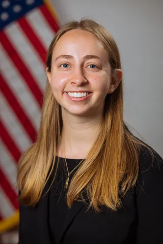 a female student with long blonde hair and a long sleeve black shirt sits in front of an American flag