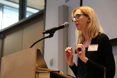 A color photo of a woman speaking at a microphone.