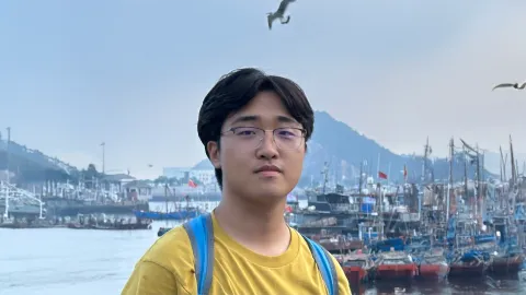 A color photo of a man with glasses in front a bay with boats.