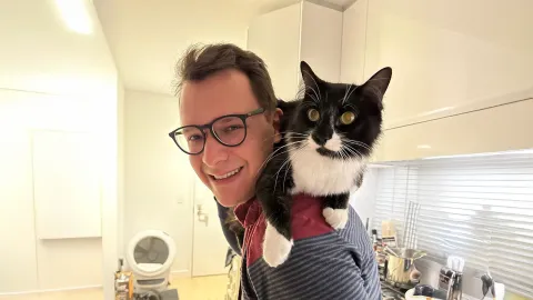 A color photo of a man with glasses smiling for a photo while a black and white cat sits on his shoulders.