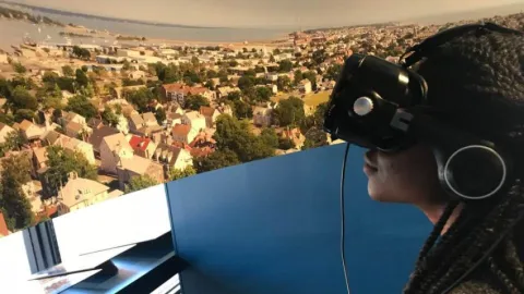 A participant wearing VR-type glasses with a visual of a sprawling suburb in front of them.