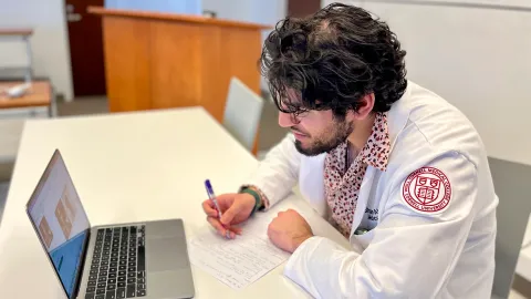 A medical student in a lab coat takes notes in front of a laptop