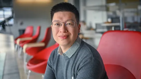 a male with dark brown hair and glasses waering a dark gray sweater with collar smiles as he sits on a red chair