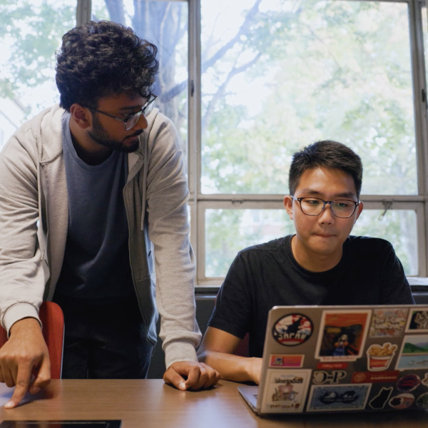 two students (one standing, one sitting) look at computer laptop