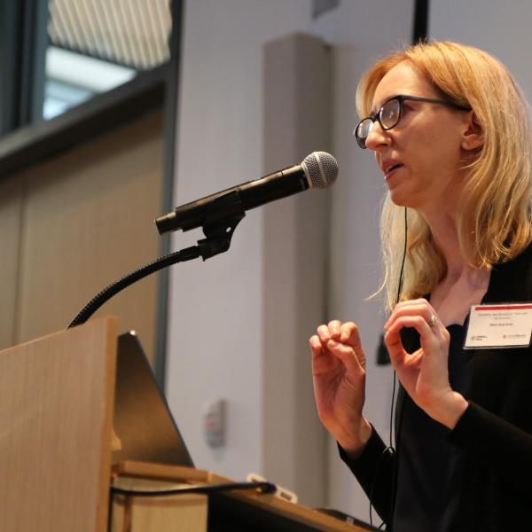 A color photo of a woman speaking at a microphone.