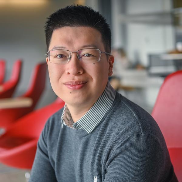a male with dark brown hair and glasses waering a dark gray sweater with collar smiles as he sits on a red chair