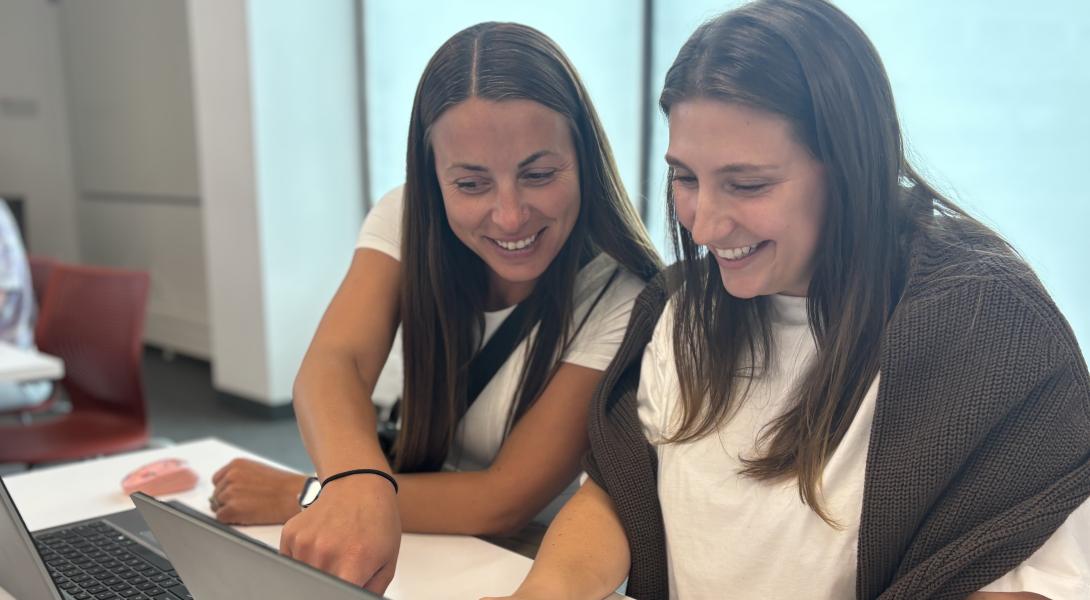 Two females with long brown hair work on a laptop together
