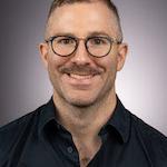 a man with glasses and light hair wearing a black button down shirt