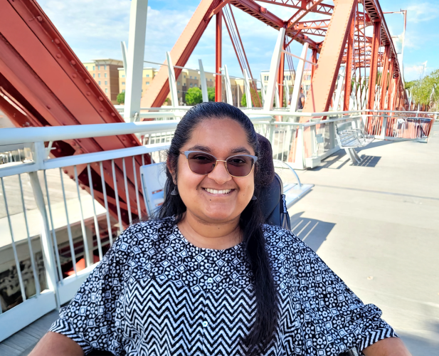 A color photo of a woman sitting outside wearing sunglasses.