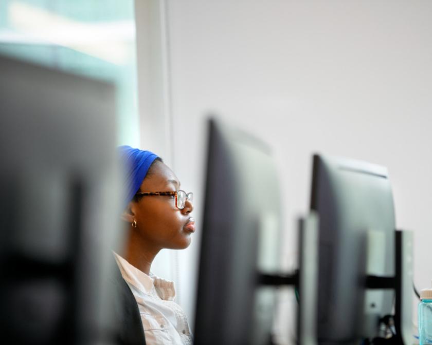 a black female student wearing a bright blue headpiece sits in between black computer monitors