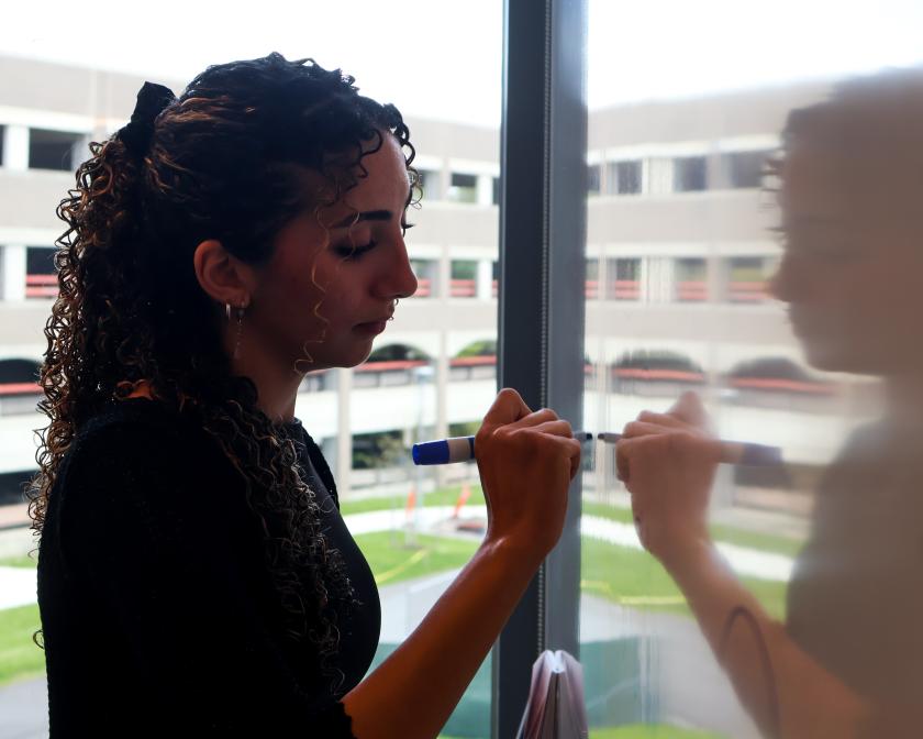 a female student with tan skin and long curly dark hair writes on white board with the reflection of her