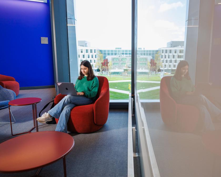 a young female student with brown hair and a green sweater sits on a red chair with her laptopr 