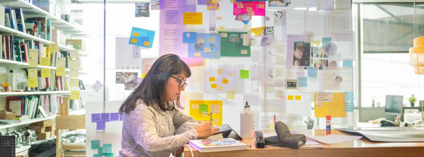A color photo of a woman working at a desk, surrounded by notes.