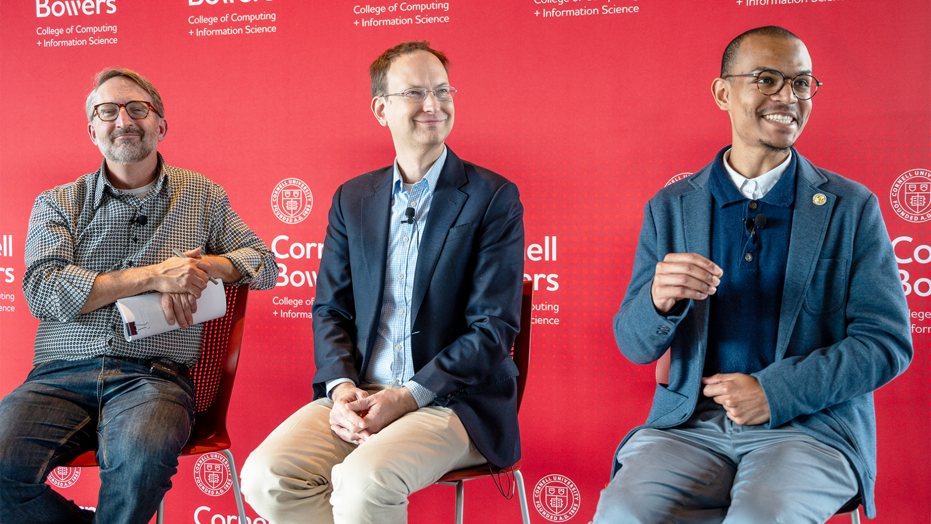 A color photo showing 3 men sitting on a stools, on a stage.